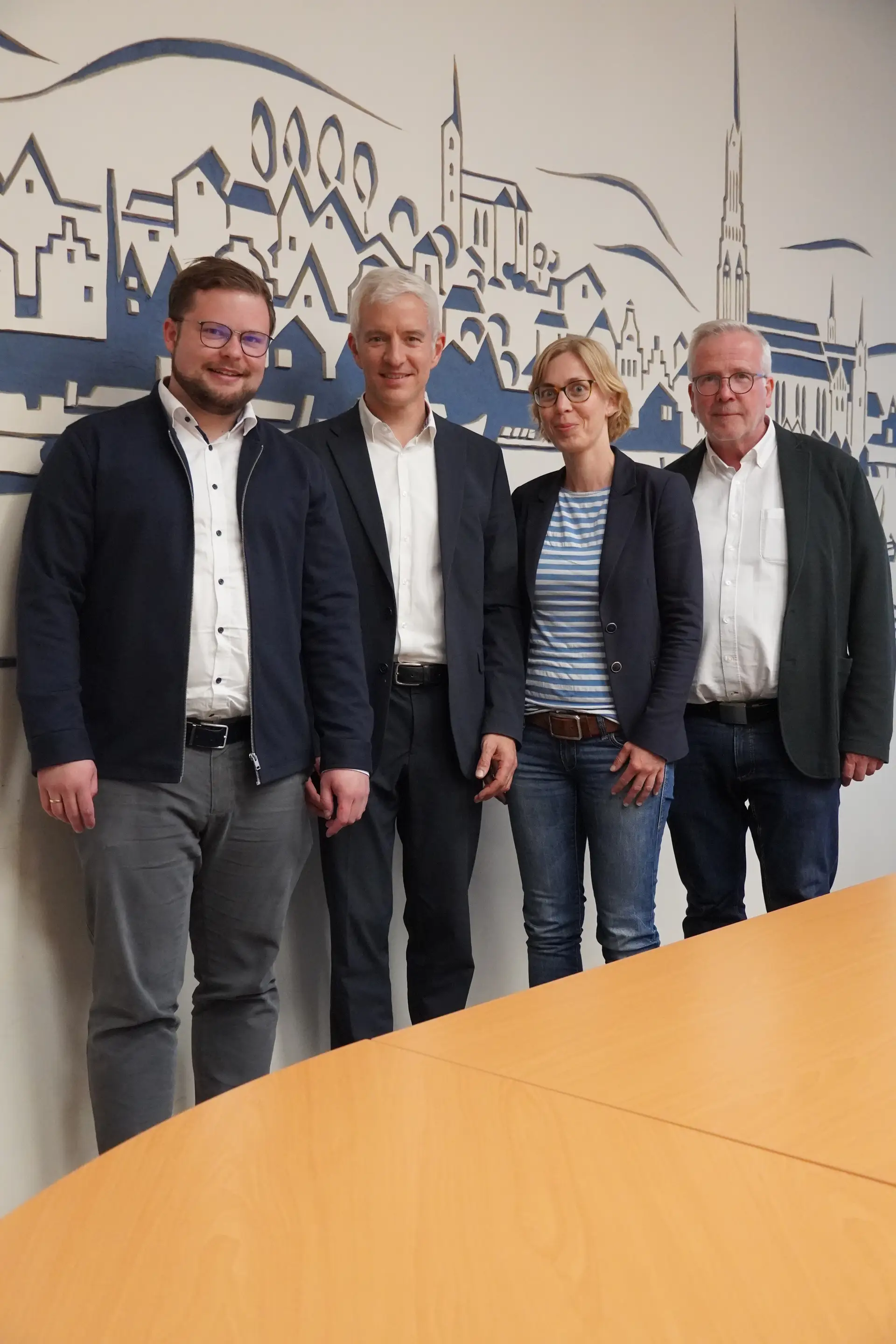 Vier Personen halten eine Urkunde/Dokumente im Rahmen einer Beurkundung; sie stehen nebeneinander vor einer Beschriftung bzw. Unterlagen im Hintergrund.